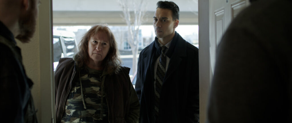 A woman in a camouflage shirt and a man in a suit stand at a doorway, looking inside with serious expressions.