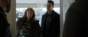 A woman in a camouflage shirt and a man in a suit stand at a doorway, looking inside with serious expressions.
