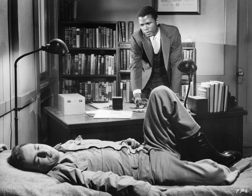 A man in a suit stands behind a desk with books, watching another man in military attire who is lying on a bed in an office filled with bookshelves.