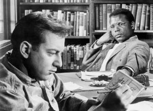 Two individuals are depicted in a scholarly setting with bookshelves in the background. A man in the foreground is writing while another man in the background looks on thoughtfully.