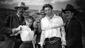 A group of people in Western attire stand outdoors with a man in a white shirt tied up with rope at the center. Mountains and sky are visible in the background.