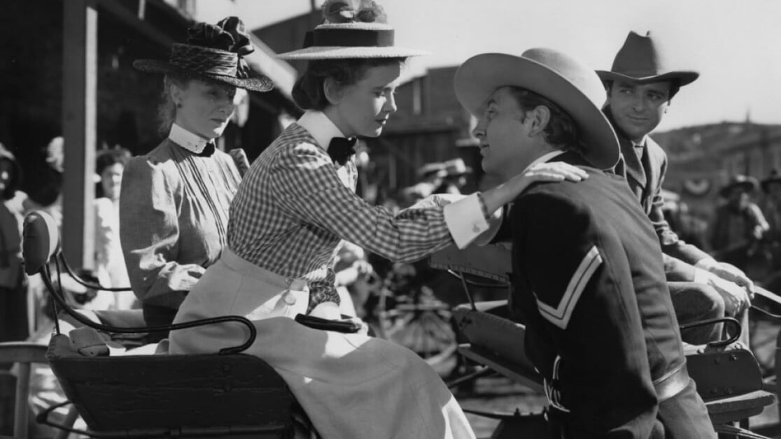 Four people in historical attire are interacting near a horse-drawn carriage. A seated woman gently touches the shoulder of a kneeling man. Another woman and a man, both wearing hats, look on.