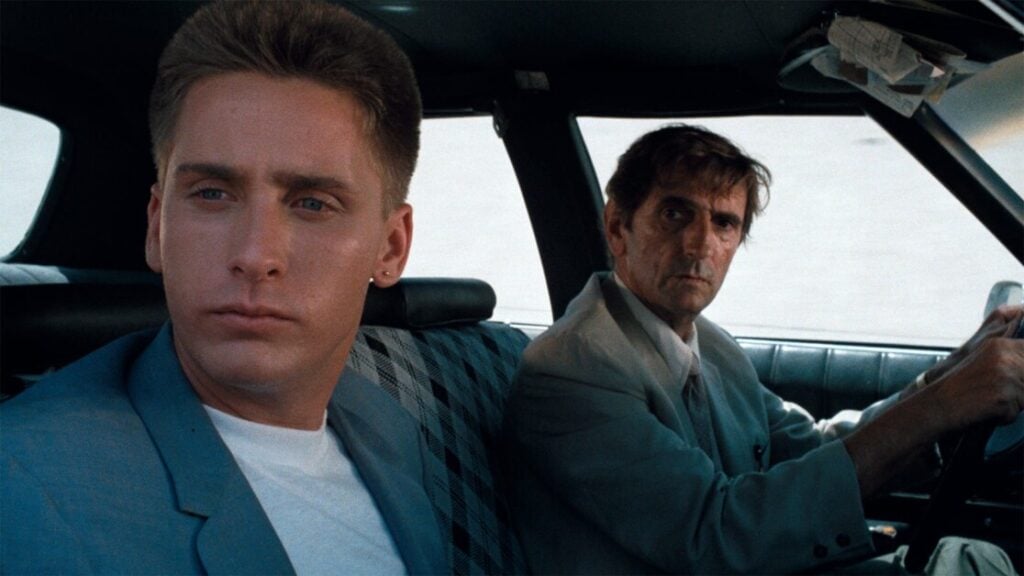 Two men sit in a car, one in the driver's seat wearing a suit, and the other in the back seat, also wearing a suit. Both appear serious and are looking in different directions.