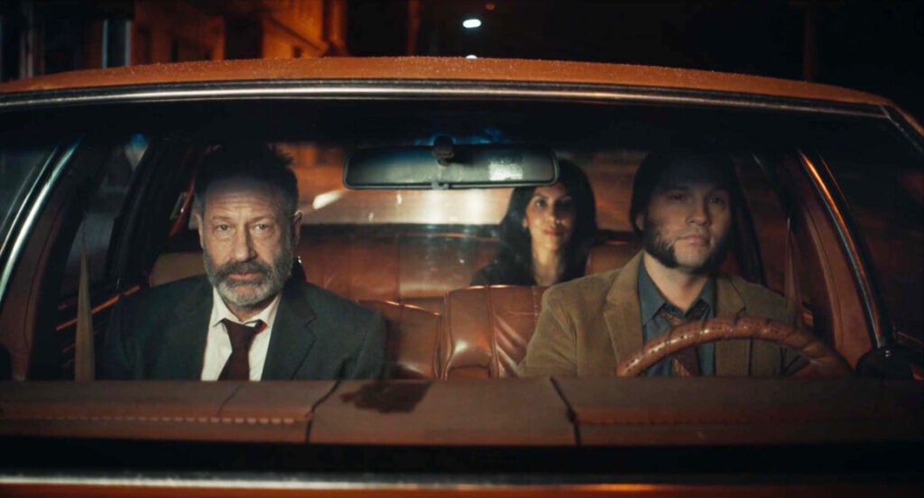 Three people sit in an old car parked on a dimly lit street at night; two men in front, one wearing a suit and tie, and a woman in the backseat. The atmosphere appears tense.