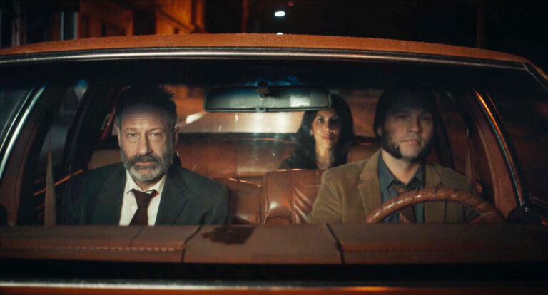 ‘Reverse The Curse’ Review – Solid Dramedy Swings For The Heartstrings Three people sit in an old car parked on a dimly lit street at night; two men in front, one wearing a suit and tie, and a woman in the backseat. The atmosphere appears tense.