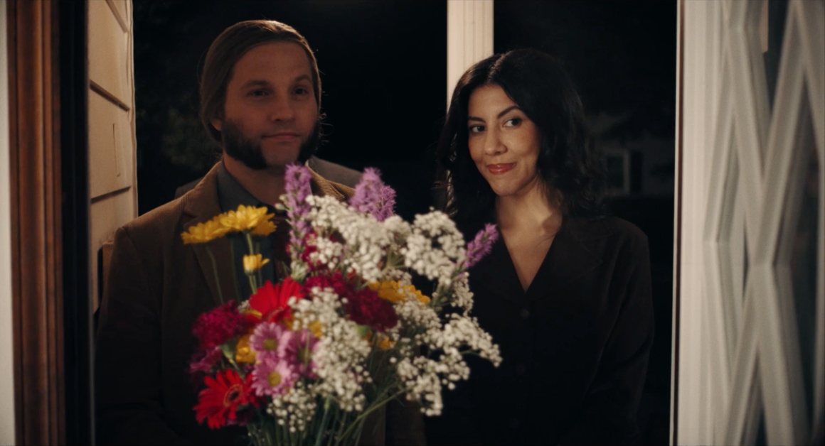 A man and a woman stand at a doorway holding a colorful bouquet of flowers, both smiling slightly, at night.