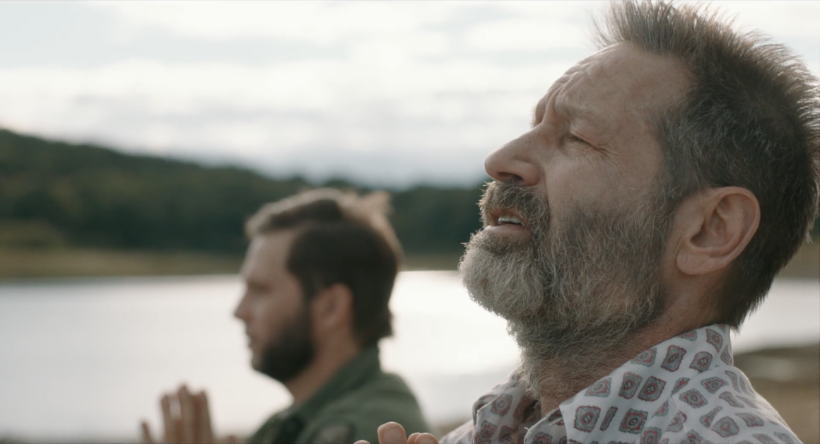 Two men are seen facing right with their eyes closed and hands clasped together, appearing to be in prayer or deep concentration, with a body of water and a forested background behind them.