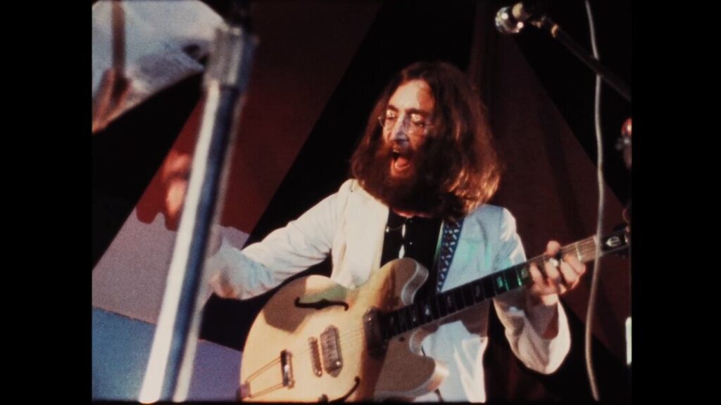 Man with long hair and glasses playing an electric guitar on stage while singing, wearing a white jacket.