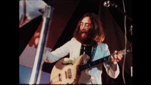 Man with long hair and glasses playing an electric guitar on stage while singing, wearing a white jacket.