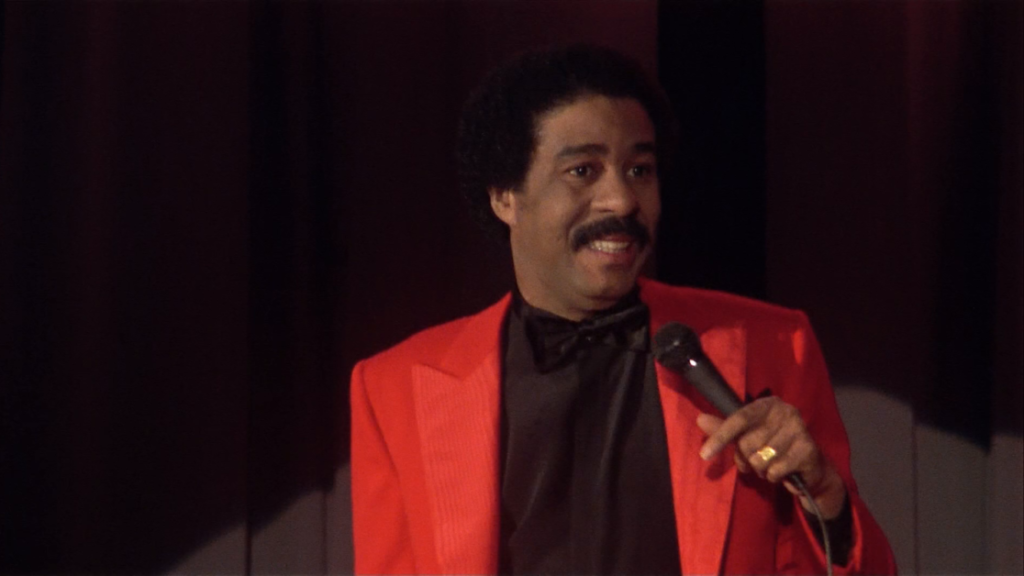 A person in a red suit and black shirt holds a microphone while smiling.