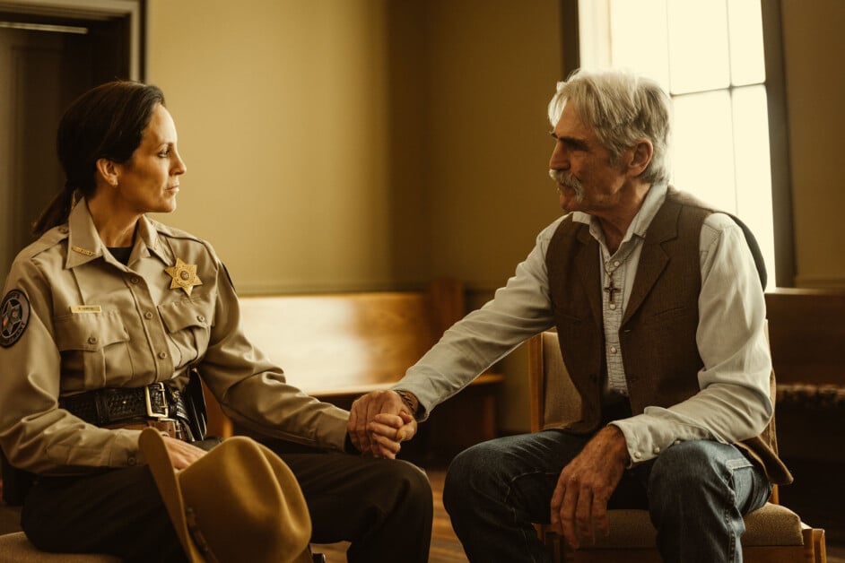 A uniformed officer and an elderly man sit facing each other in a dimly lit room, holding hands as they engage in a serious conversation.