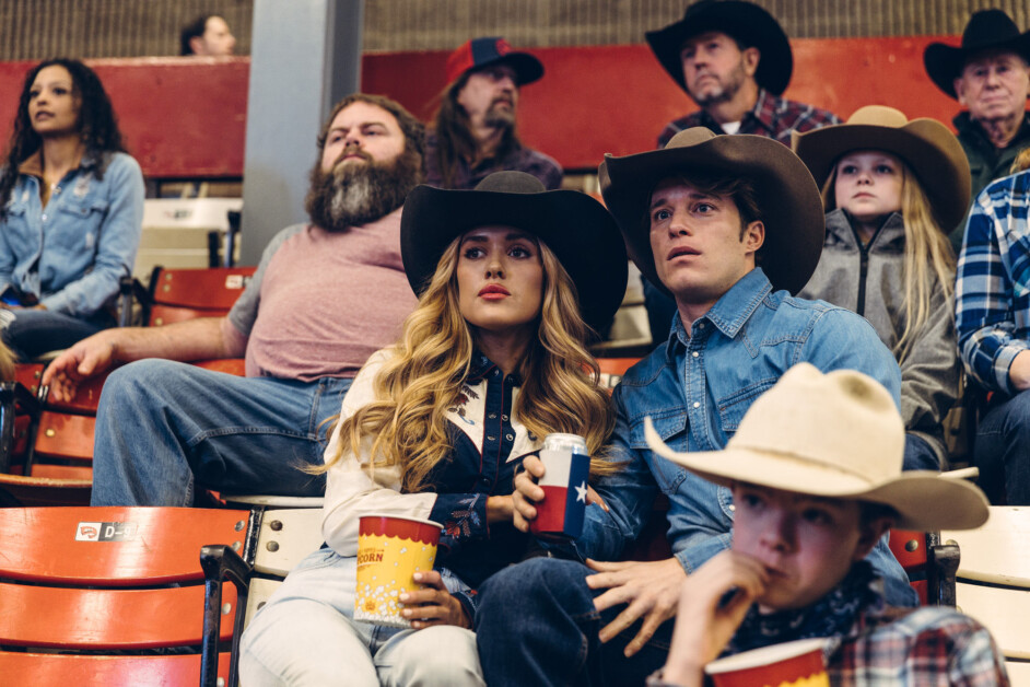 A group of people, wearing cowboy hats and casual clothing, sit in stadium-style seats, intently watching an event. Two individuals in the foreground hold popcorn and drinks.