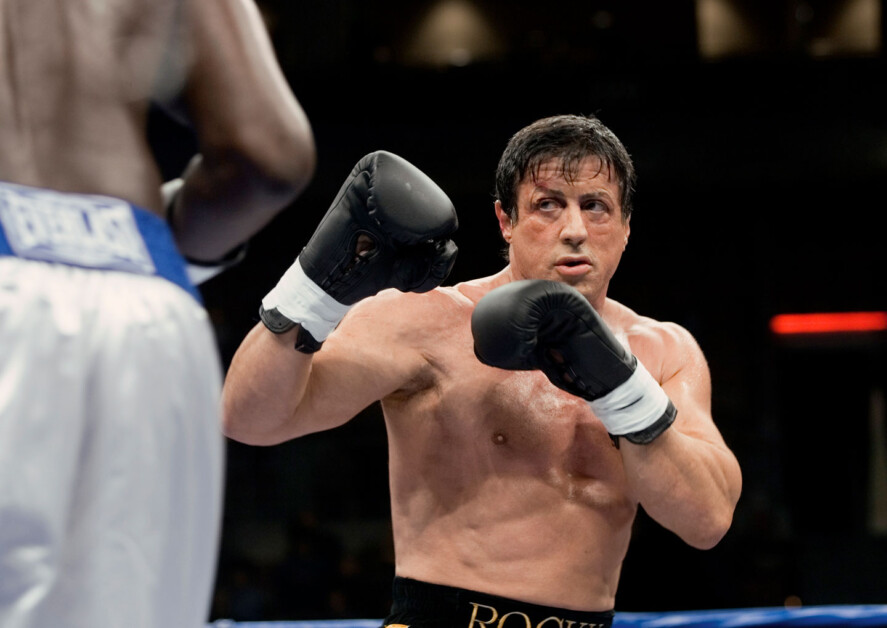 A boxer in black gloves and shorts prepares to strike, while another boxer in white shorts stands opposite in a boxing ring.