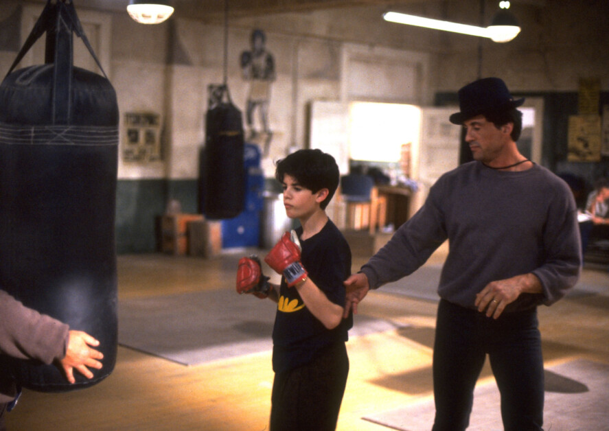A young boy wearing red boxing gloves stands in a boxing gym with a man in a hat and sweatshirt beside him. They appear to be in a training session with a punching bag in the background.