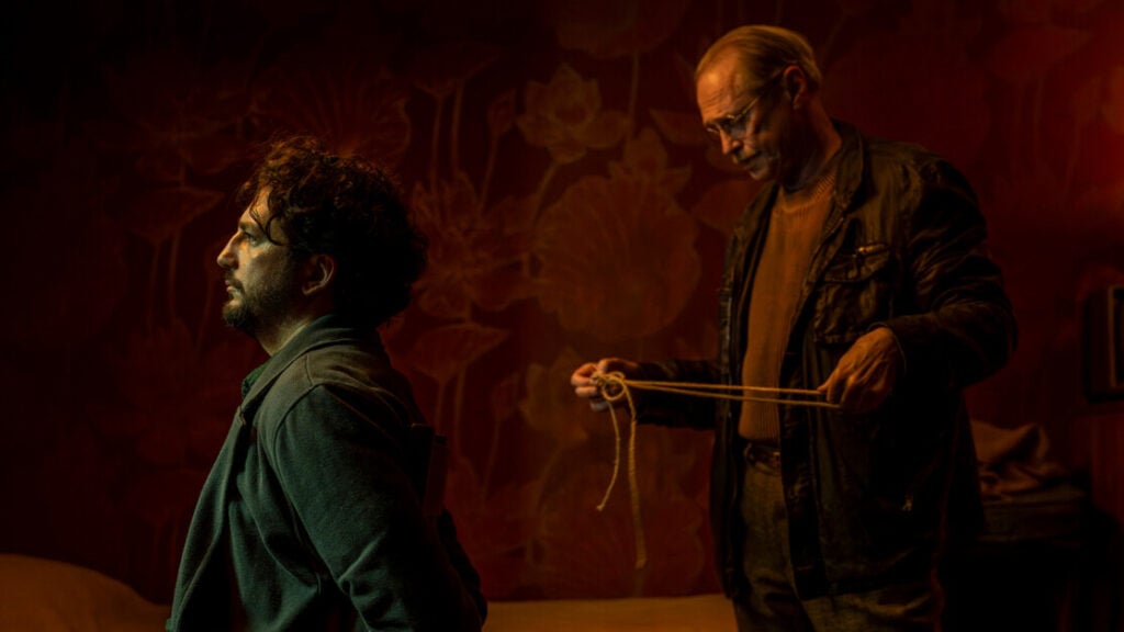 Two men in a dimly lit room. One is seated and appears concerned, while the other stands behind holding a rope, focused on it. The background features floral wallpaper.