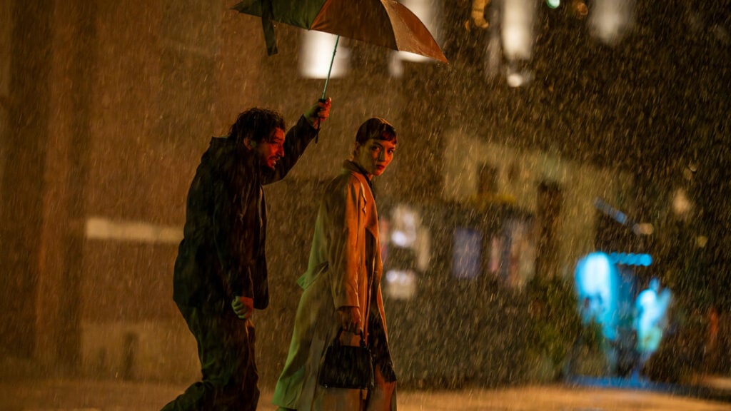 Two people walk in heavy rain, one holding an umbrella to shield both. Their expressions indicate urgency. They are in an urban setting with blurred lights in the background.