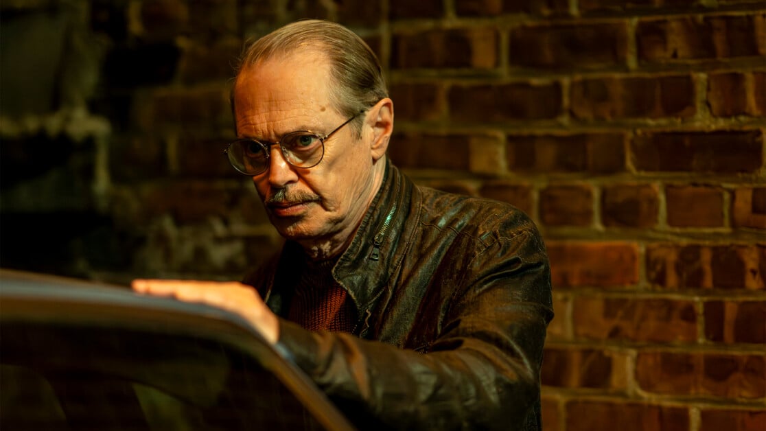 A man with glasses and a mustache, wearing a leather jacket, stands by a car against a brick wall.