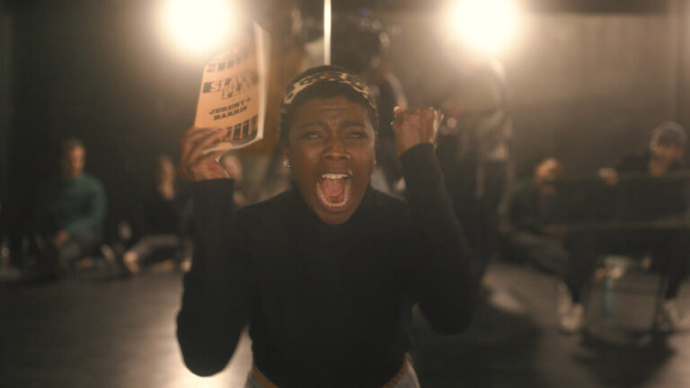 A person stands in a dimly lit room, holding a book and shouting with an intense expression. Other individuals are seen in the background.