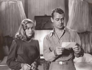 A woman and a man in 1940s attire are indoors, with the man holding and looking at a piece of paper while the woman looks on.
