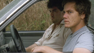 Two men sitting in the front seat of a vehicle; the driver looks directly at the camera with a serious expression, while the passenger looks off to the side.