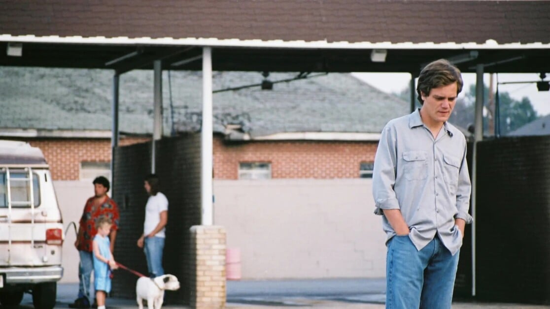 A man in a gray shirt and jeans stands with his head down under a carport with two people and a dog in the background.