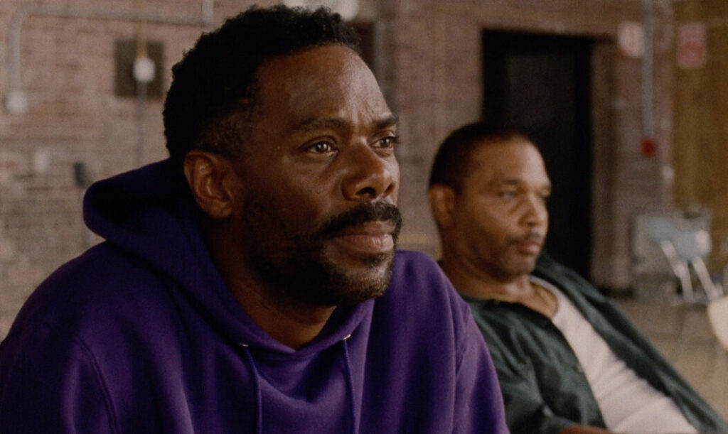 Two men sit indoors, with one in the foreground wearing a purple hoodie and looking forward, while the other is in the background seated and looking slightly to the side.