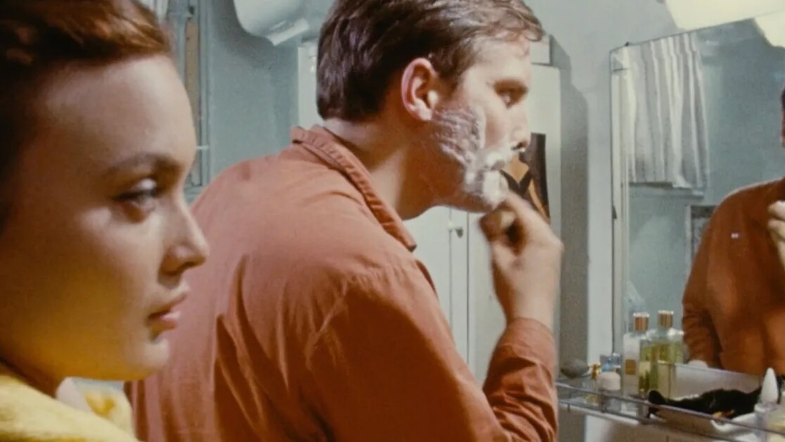 A man shaves in front of a bathroom mirror while a woman looks on from the side.