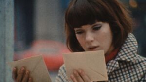 ‘Six In Paris’ Blu-Ray Review – Terrific Anthology Film Gives You A Taste Of The French Masters A person with short brown hair and wearing a checkered jacket looks intently at two envelopes.