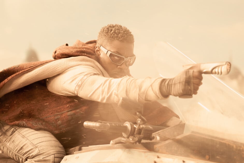A person in futuristic attire rides a high-speed vehicle, extended arm pointing a gun forward. Dust and debris surround them, suggesting a high-intensity situation.