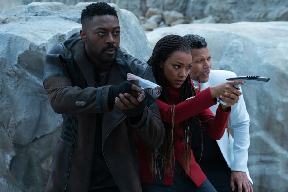 Three individuals in futuristic attire holding weapons and pointing forward. They appear focused and alert while standing against a rocky background.