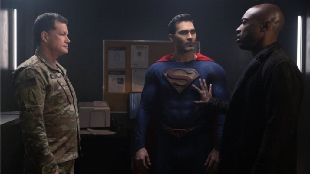 Three men converse in a dimly lit room. One wears a military uniform, another a superhero costume with an 'S' emblem, and the third is dressed in black, placing a hand on the superhero's chest.