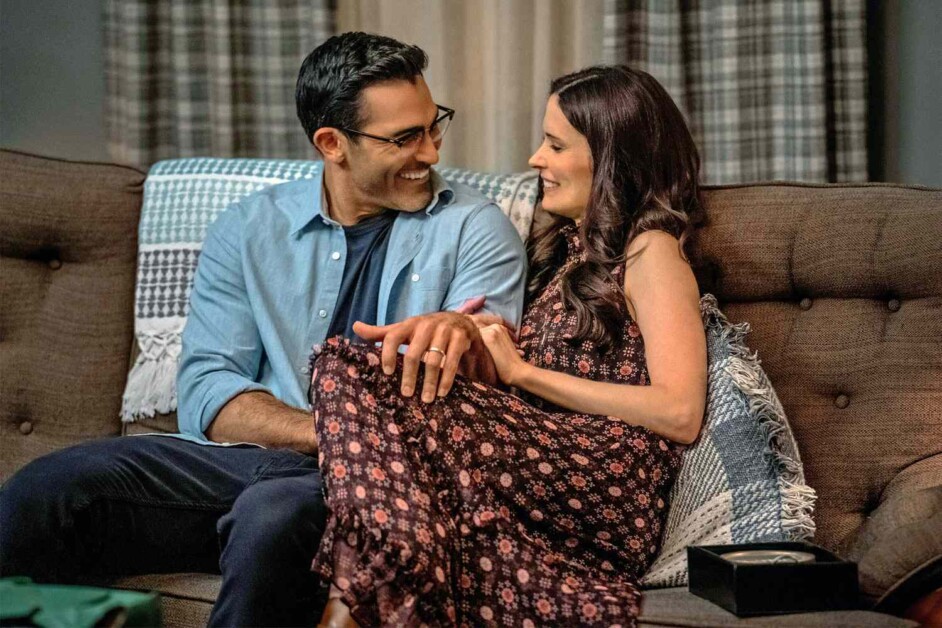 A man and woman sit close together on a couch, smiling and holding hands. The woman wears a floral dress, and the man wears glasses and a blue shirt.