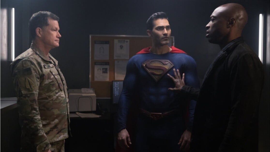 Three men converse in a dimly lit room. One wears a military uniform, another a superhero costume with an 'S' emblem, and the third is dressed in black, placing a hand on the superhero's chest.