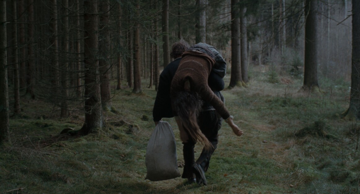 A person is carrying another person over their shoulder while walking through a forest, holding a sack in one hand.