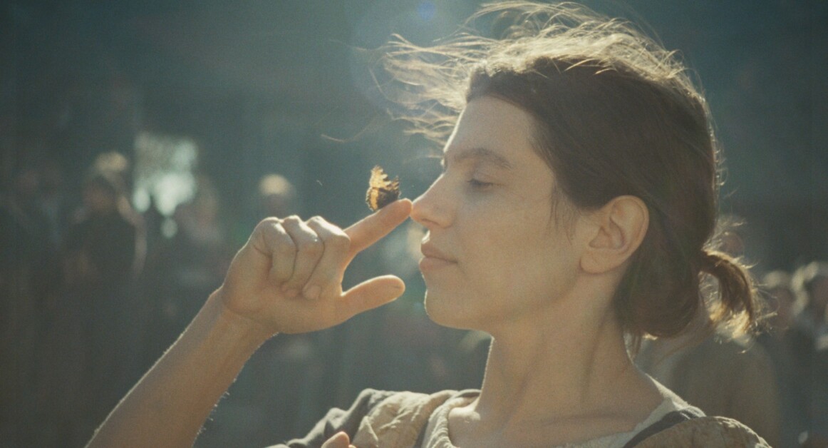 A person with a butterfly perched on their nose holds their index finger near the butterfly while standing outdoors.