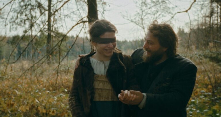 A woman with a blindfold and a man with a beard walk hand in hand through a forest. Both are smiling and dressed in rustic clothing.