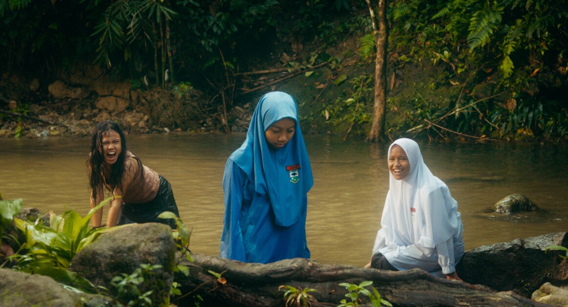 Three girls are at a river; two are standing in the water, one in a blue hijab looks down while another in a white hijab smiles, and the third girl, in casual clothes, laughs while playing in the water.