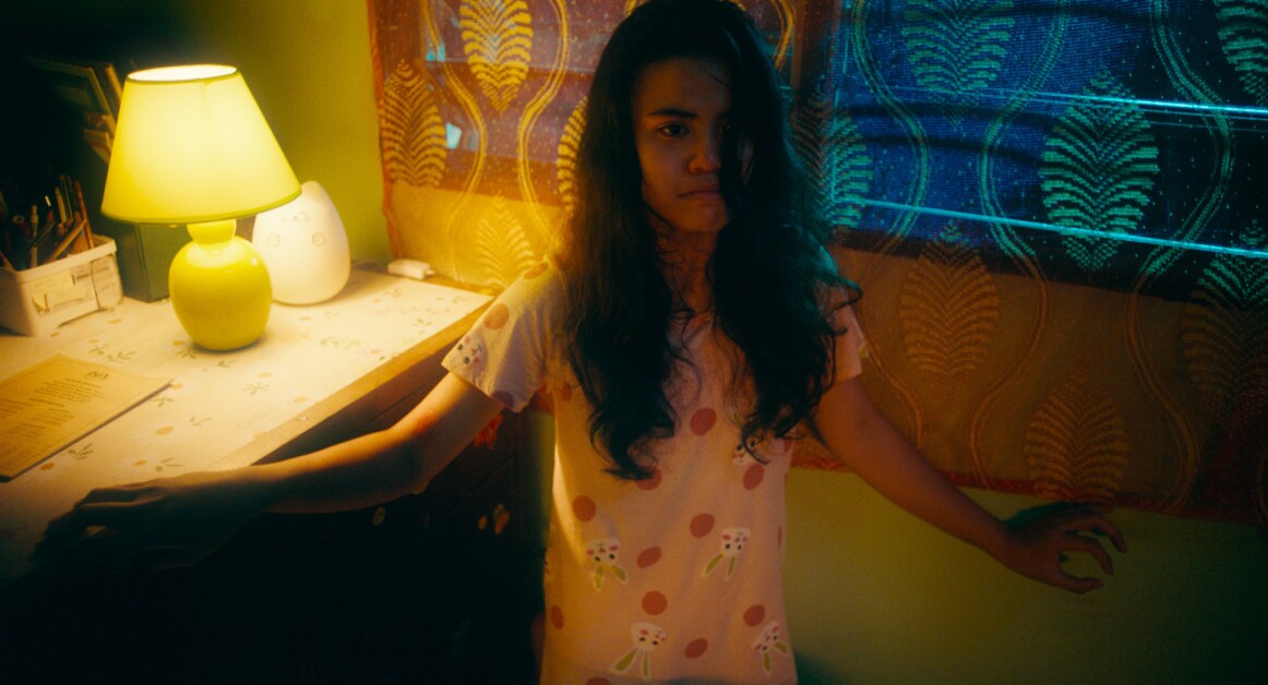 A person with long hair stands in a dimly lit room next to a lamp and window with patterned curtains. They appear to be looking forward with a slightly tense expression.