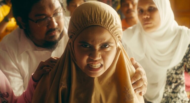 A girl wearing a beige hijab appears angry, with two adults, one in a white shirt and one in a white hijab, on either side of her. Several other people are visible in the background.