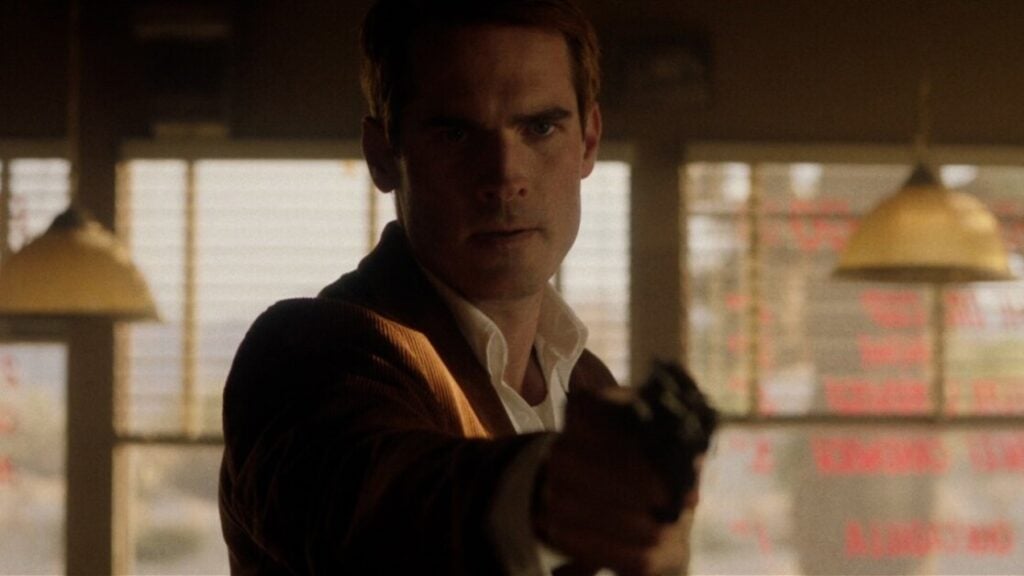 A man stands indoors, pointing a gun forward with a serious expression. Blurred background suggests a well-lit room with hanging lights and window blinds.