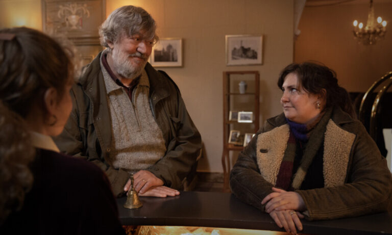 ‘Treasure’ Review – Aspiring Tearjerker Is A Flurry Of Shortcomings Two people warmly dressed for winter stand at a counter, engaging with a third person. The background features framed pictures and a chandelier.
