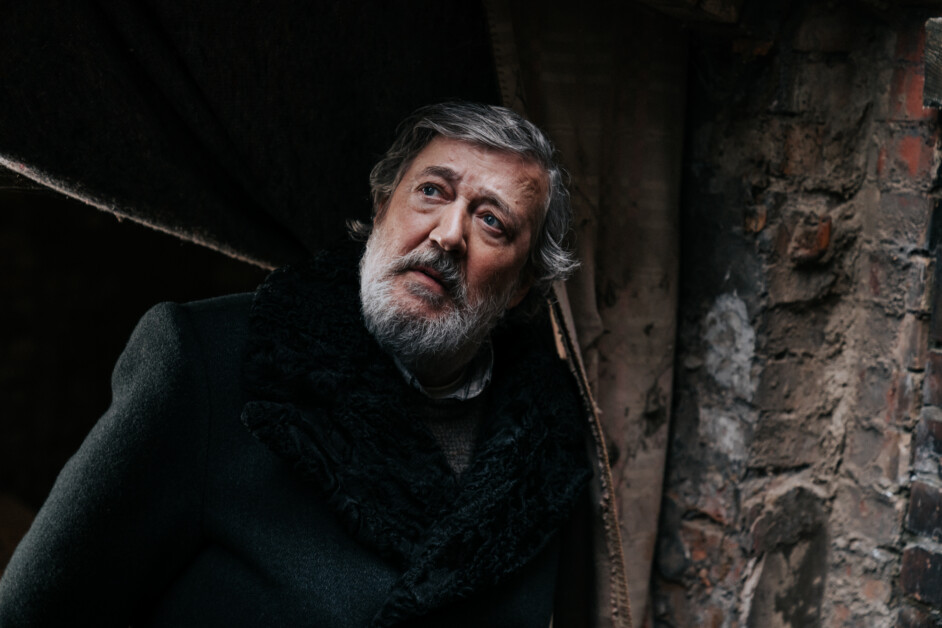 An older man with a gray beard and dark coat looks upward in a dimly lit, rustic setting with brick and worn fabric in the background.