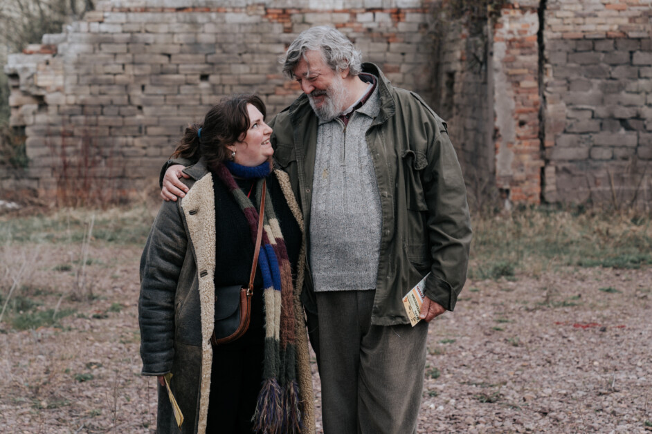 Two people stand outside near a brick wall, wearing winter clothes. The older person has their arm around the younger, and they're both smiling. The older person holds a small book or pamphlet.