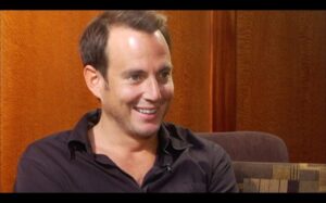 ‘The Arrested Development Documentary Project’ Blu-Ray Review – A Love Letter To A Beloved Series A man with short brown hair and stubble is sitting indoors, smiling, and wearing a dark shirt. The background features a textured orange wall and a patterned cushion.