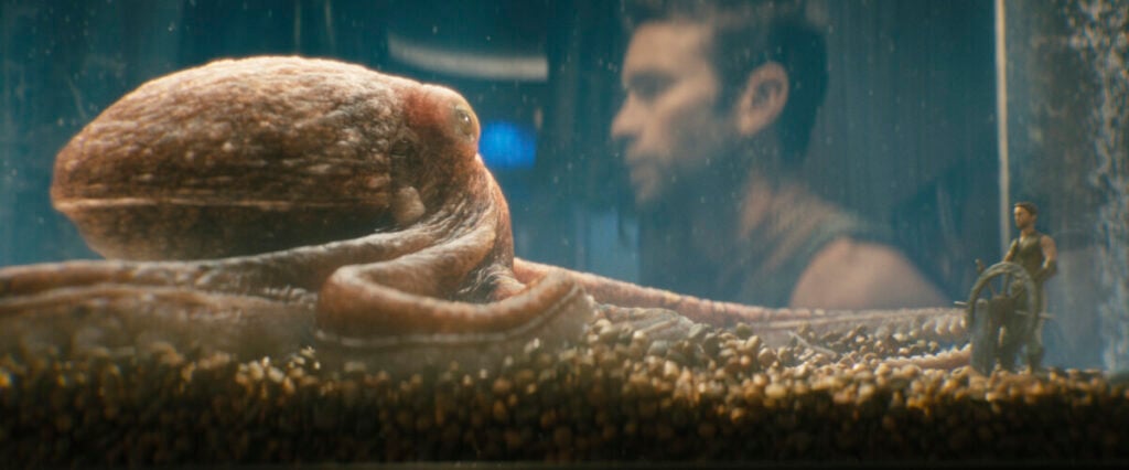 An octopus inside a tank is observed by a blurred human figure in the background, with another small figure guiding a ship wheel in the foreground.