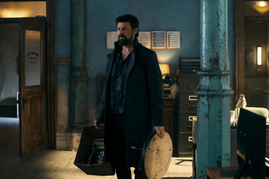 A man with a beard and dark coat stands in a dimly lit room holding a box and a round object. A desk and office equipment are visible in the background.