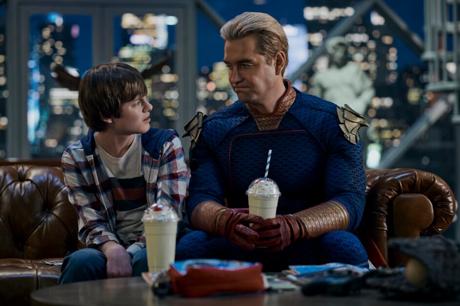 A child and an adult in a superhero costume sit together on a couch with milkshakes in front of them. The adult looks at the child, and they are in a room with a cityscape view in the background.