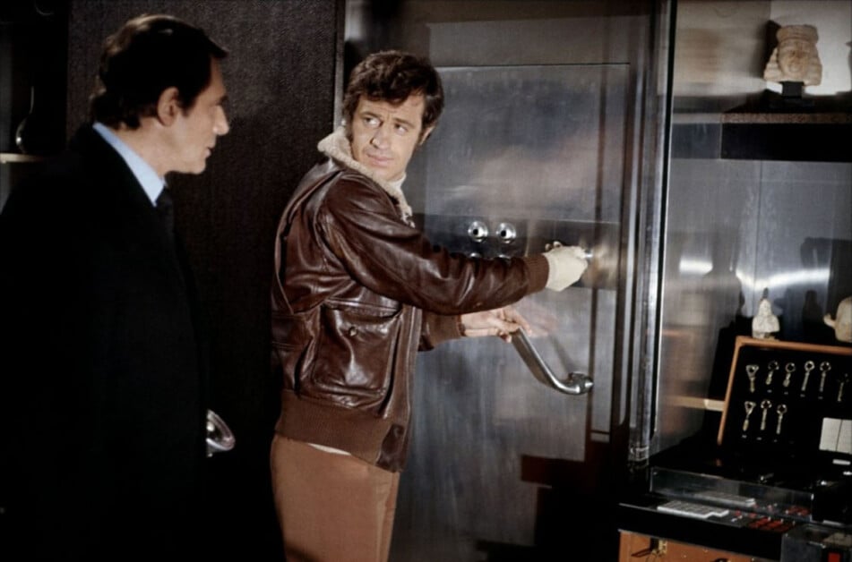 Two men are in a room with a large metallic safe; one man in a leather jacket is attempting to open it while the other man, dressed in a suit, watches.