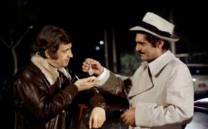 ‘The Burglars’ Blu-Ray Review – Thrilling Caper Offers Action That Can’t Be Beat Two men are engaged in an interaction at night; one man in a leather jacket is holding a small object while the other, in a trench coat and hat, hands it over. A car is partially visible behind them.