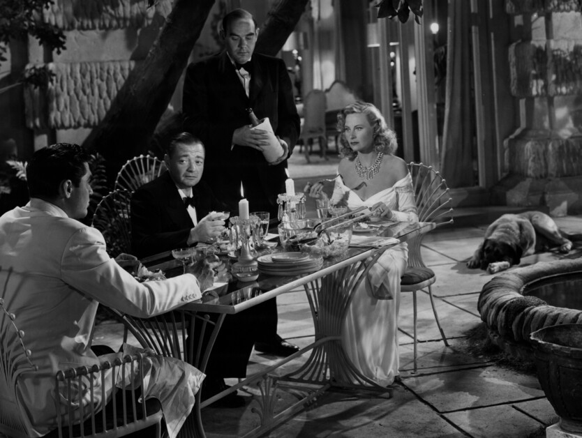 A group of four people dressed formally are seated around a dining table outdoors at night. A waiter stands behind them, and a dog is lying on the ground next to the table.